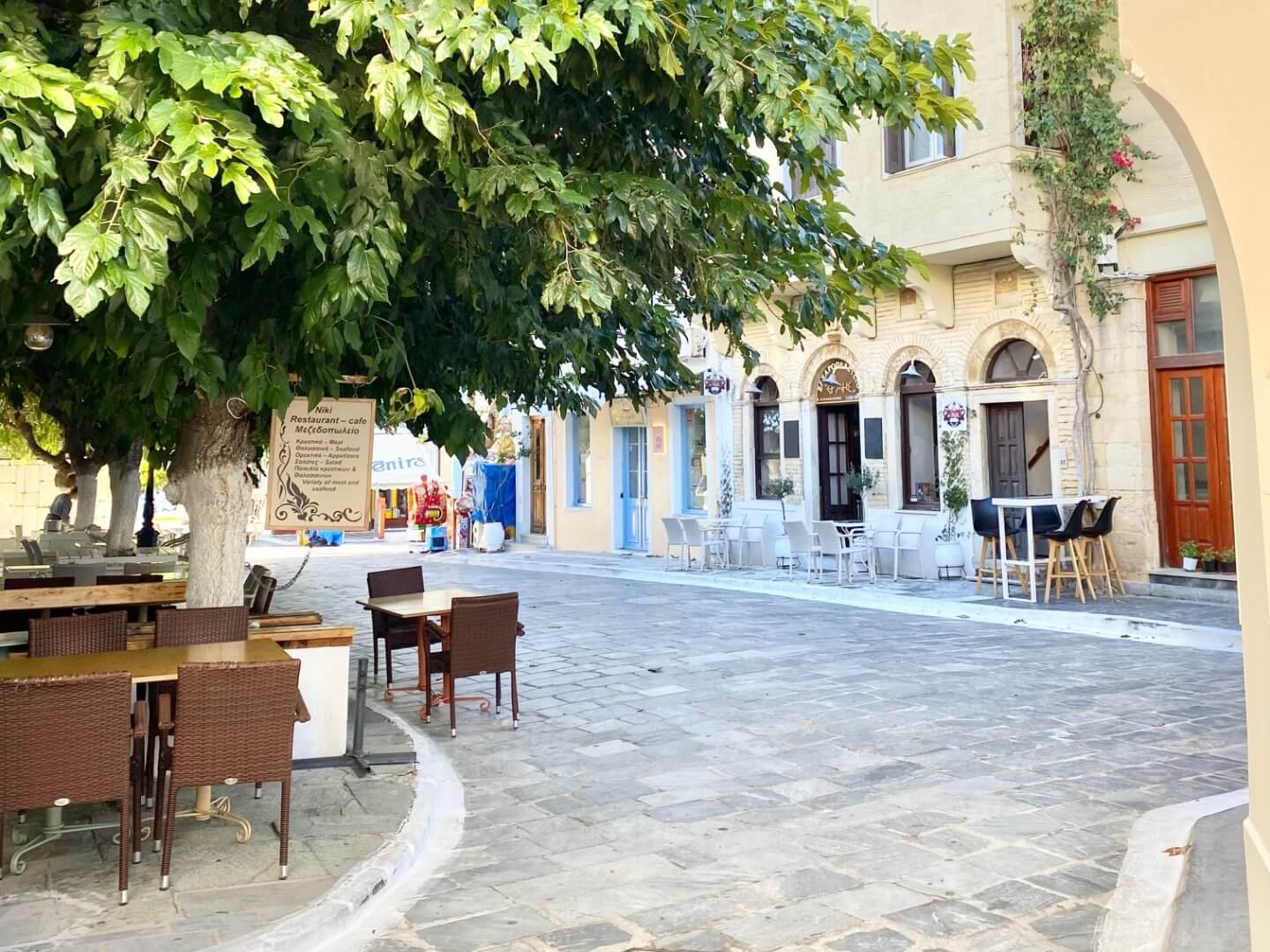 Square with cafe tables and chairs on the street and under plane tree - example of Where To Eat In Andros