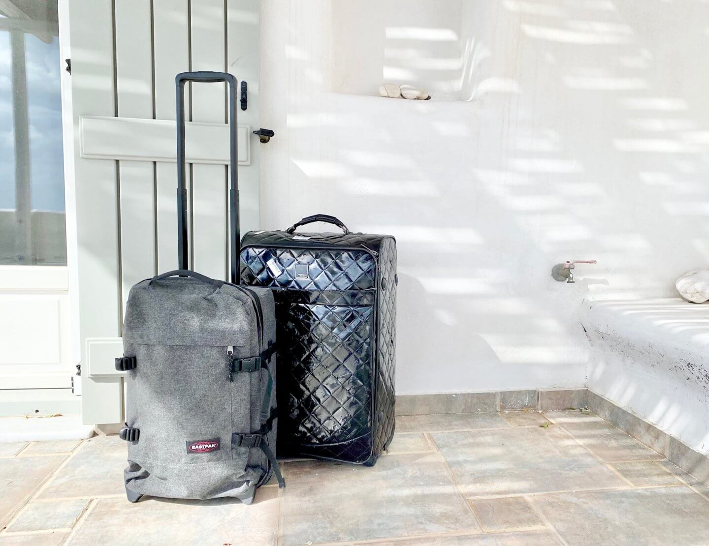 Black shiny quilted suitcase and smaller grey denim suitcase with handle up on whitewashed terrace