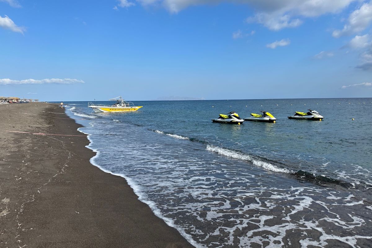 Yellow boat and three jet skis in the water on Perissa black sand beach